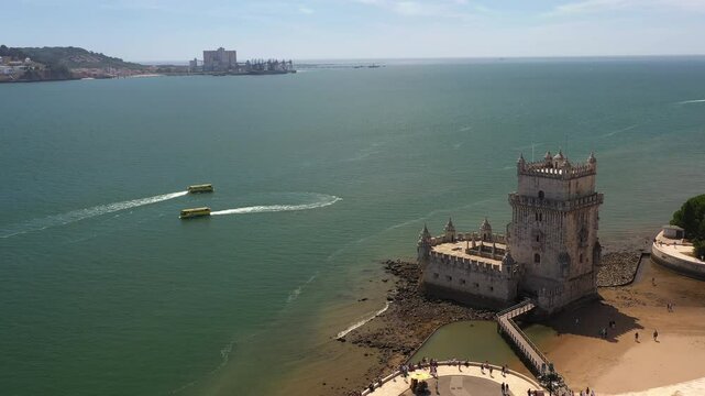 Aerial view of Belem Tower on Tagus River with waterfront in background. Amphibious buses moves in circular pattern creating wakes in water