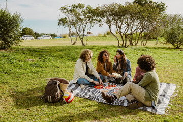 A multinational group of young people is sitting on a blanket in a park on a sunny day, enjoying a...