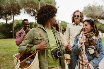 A multiracial group of young friends walks together in a park. An Asian woman holds a small dog in...