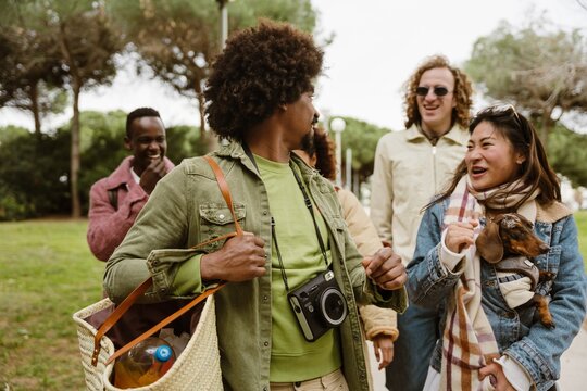 A multiracial group of young friends walks together in a park. An Asian woman holds a small dog in her jacket while talking to a man carrying a bag and camera. Others laugh and walk behind them.