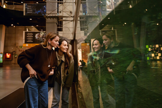 Two adult women at an aquarium exhibit stand close to fish through the glass. Their reflections are visible, and they smile and enjoy the experience in a public space.