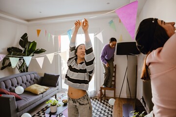 Three young, Asian and brown-skinned female friends decorate the living room for a party together,...