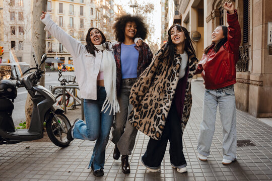 Four young, diverse female friends in fashionable outfits have fun together while strolling down a street. A concept of fashion, friendship, girlhood, and youth.
