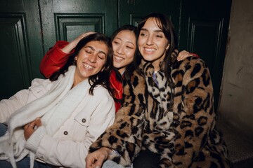 Plakat A multinational group of young women sitting closely together at a girl's party, laughing and smiling joyfully, dressed in casual and stylish jackets, enjoying a cozy and cheerful evening outdoors.