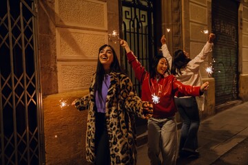 In a dimly lit street at night, three women dance in a row, laughing and waving sparklers in the...