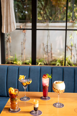 A vibrant lineup of five differently colored cocktails in shooter, goblet, collins, martini, and highball glasses on a brown table, sunlight pouring in with plants and sofa in the background.   