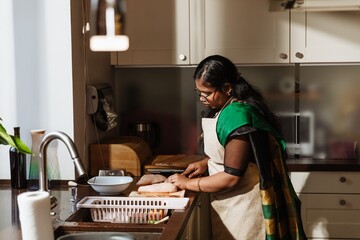 An adult Indian woman in a green and black sari and apron prepares raw meat in a modern kitchen. She cuts food on a board at the counter near the sink.