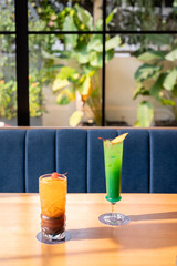 Brown and green cocktails in highball and milkshake glasses on a sunlit table, with blurred sofa, window, and plants in the background of a cozy restaurant.