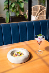 Red ruby dessert and gulab jamun with rabri placed on a restaurant table bathed in sunlight, with a blurred sofa and green plants in the background.