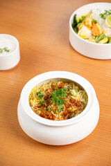 Fragrant biryani with vegetables, paneer, mint, coriander, and saffron served in a white pot, paired with curd and a blurred side dish on a brown restaurant table.      