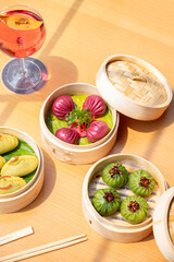 Top view of Wild Mushroom Dim Sum, Cantonese Prawn Dumpling, and Crystal Vegetable Dumpling served with dipping sauces and chopsticks, paired with a red cocktail on a sunlit restaurant table.   