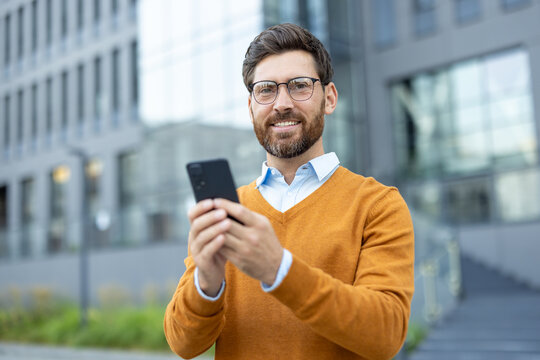 A smiling businessman, holding his phone, stands confidently outside in front of a modern building, showcasing technology.