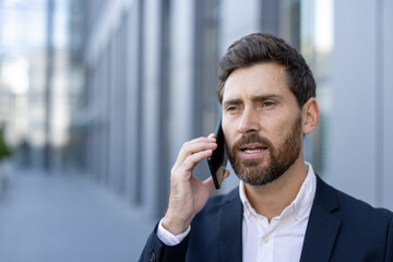 A handsome businessman, wearing a suit, uses his phone while standing in front of an office...