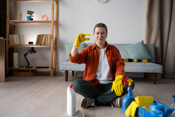 Housekeeping job concept. Young woman in casual clothes sitting on floor with cleaning supplies, washing and doing homework cleaning routine looking at camera showing small gesture