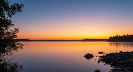 Obraz premium Serene Sunrise over Calm Lake with Silhouetted Trees and Rocks