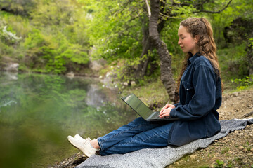 Serious young woman with curly hair looking at laptop computer screen distance learn remote work rest relax in daylight sit in plaid in forest near a lake. Digital nomad lifestyle outdoors in nature
