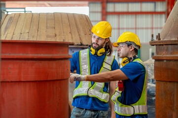 Two engineers in hard hats and safety vests discuss a large metal structure in a factory.