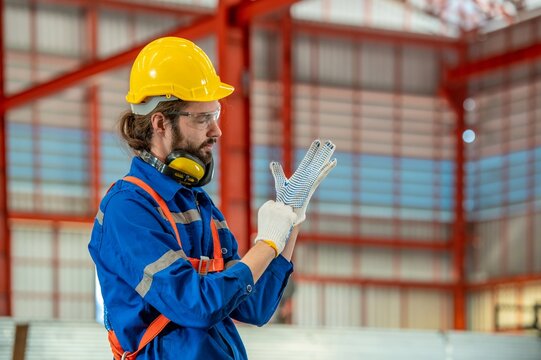 A man wearing a hard hat and safety vest is putting on his gloves in a factory.