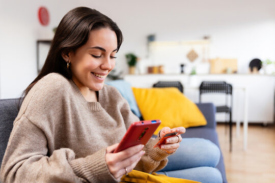 Young happy woman making online payment using mobile phone and credit card, enjoying convenient and secure e-commerce experience from home