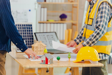 Architects and solar engineers discuss home design in office using model homes, solar panels, and architects' design documents. Concepts about home construction and solar power.