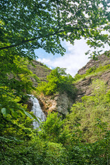 A hidden waterfall cascading down rocky cliffs, framed by lush green trees in a remote forest valley. Scenic natural spot in the Georgian mountains