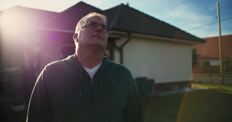Man gazing upward outside residential house, wearing glasses, peaceful moment in suburban garden,...
