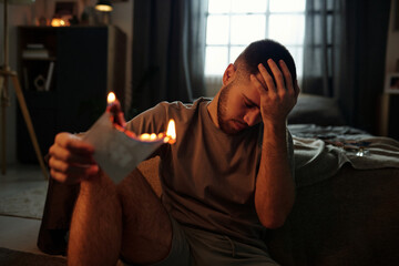 Young man sitting in dimly lit room, holding burning paper in hand, appearing deep in thought with...