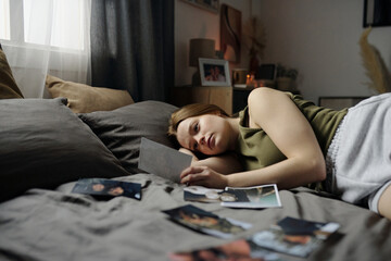 Woman lying on bed in cozy room, holding and looking at photograph while surrounded by other...