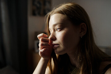Woman sitting indoors resting her chin on hand and gazing out window reflecting on something while...