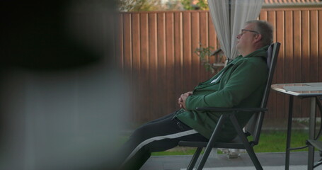 Man reclining on patio chair with arms crossed, resting in backyard with wooden fence and greenery, enjoying quiet relaxation and outdoor serenity