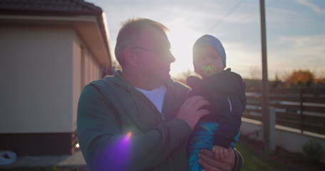 Father holding baby with pacifier in backyard, gently adjusting child’s outfit, showcasing...