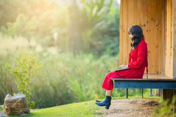 Beautiful asian woman is sitting relaxed with laptop at terrace in the greenery. Beautiful morning light in garden with green grass field. Free space for advert