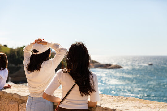 Deux jeunes femmes de dos avec cheveux au vent fl&acirc;nant et regardant la mer au soleil au premier plan et ciel bleu lumineux avec mer brillante en arri&egrave;re plan dans une ambiance de vacances