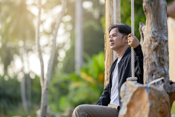 Asian man enjoys a peaceful nature on a wooden swing overlooking the vast scenic landscape. Bright sunny day