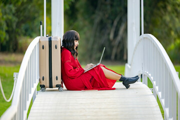 Young woman wear summer casual clothes sit near suitcase using laptop for online search for holiday destination or discount offers on travel while sitting on bridge relaxing outdoors feeling happiness