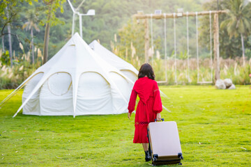 Back view young woman wear summer casual clothes walk go with suitcase camping leisure and White tent with green grass and trees in luxury camping. Tourism relax and chill summer holiday.