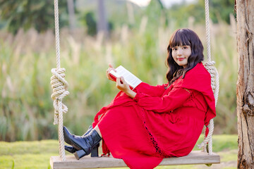 Beautiful Asian woman feeling happy smiling and looking to camera while relax sitting on a swing at flowering field and reading book.