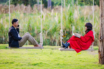 Weekends getaway. Asian couple enjoying the joys of being on an outdoor wooden swing during sunset, searching sightseeings for trip on laptop, planning route and talking on cellphone.