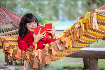 Young elegant beautiful Asian woman dressed in summer clothes smiling and  reading book lying on hammock at terrace.