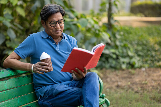 Indian Senior man reading story book while sitting on bench at park. Indian elderly gentleman enjoying the sunshine park with his reading book.