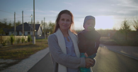 Mother carrying toddler in arms while smiling outdoors on a quiet suburban street, enjoying a peaceful moment during a sunny day near neighborhood homes