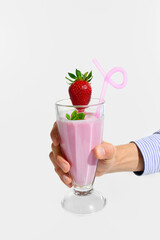 Hand holding tall glass of strawberry smoothie decorated with fresh strawberry, mint and pink curly straw against white studio background. Concept of fast food, nutrition, catering, takeaway.