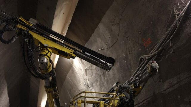 Underground tunnel drilling robotic arms extend toward concrete wall heavy cables and hydraulic tubes twist through dark confined space industrial lights highlight high-tech excavation