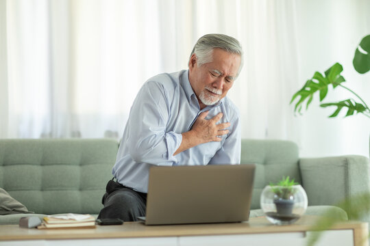 Asian elderly man experiencing chest pain while sitting on sofa with laptop in front of him, in bright living room with plants and natural light - Powered by Adobe