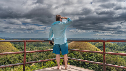 A man stands at the railing of the observation deck of the Chocolate Hills Natural Monument, looking into the distance. The view from the back. Rows of unusual rounded mountains stretch to the horizon
