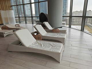 Contemporary lounge space with wicker sun loungers and soft cushions, large windows revealing city skyline, inviting relaxation and comfort in a tranquil atmosphere