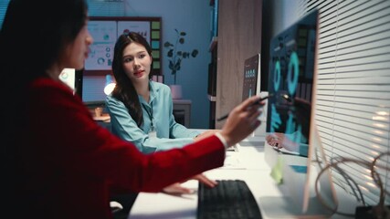 Dedicated businesswomen collaborating during late evening hours, analyzing financial charts and graphs on computer screen in modern corporate workspace - Powered by Adobe