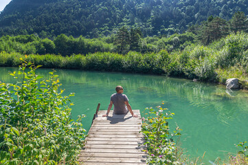 Fototapeta premium A man is sitting near a lake in the mountains. Summer vacation by the water. A man looks into the distance, sitting on the shore of the lake. The pond is overgrown with grass and shrubs.
