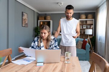 Young couple working remotely at home, analyzing documents and data, using laptop in cozy modern...