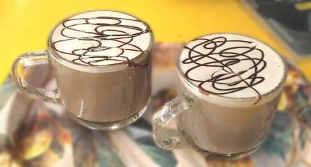 Two glass cups of hot chocolate with frothy milk and artistic chocolate syrup topping, served on a colorful tray with a vibrant background.